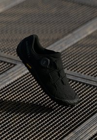 Crna atletska cipela s prozračnom mrežastom gornjom polovinom, okruglim vrhom i jedinstvenim elastičnim sistemom vezivanja. Vibram potplat s teksturiranim uzorkom.