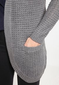 Grå stickad cardigan med en öppen vävdesign, med rundad nederkant och en sidoficka. Ärmarna är ribbade för textur.