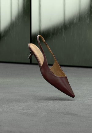 Donkerbruine leren slingback hak met puntige neus zwevend boven een grijze vloer tegen een wazige glazen raambackground.