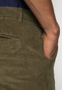 Olive green corduroy pants with raised ribbed texture, featuring a side pocket and a classic waistband design.