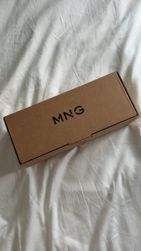 Pravokutna smeđa kartonska kutija s natpisom "MNG" ispisanim crnom bojom na vrhu, postavljena na bijelu naboranu tkaninu.