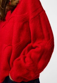 Red knitted sweater with ribbed texture, dropped shoulders, and loose fit; features visible stitches and a slightly oversized design.