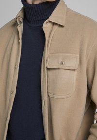 Béžová fleecová bunda se zapínáním na knoflíky, kapsou a límcem, navrstvená přes námořnický modrý žebrovaný rolák. Měkká textura.