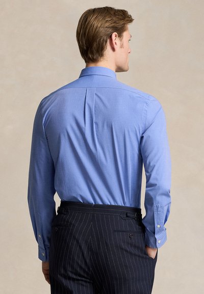 Hátulról látható férfi, aki világoskék hosszú ujjú ingben, fekete csíkos nadrágba betűrve áll.