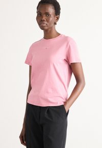 Kortærmet lyserød bomulds T-shirt med rund hals. Har et lille logo på brystet og en afslappet pasform, kombineret med sorte bukser.