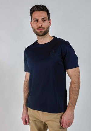 Homme aux cheveux courts et foncés avec une barbe, portant un t-shirt bleu marine et un pantalon beige, debout devant un fond uni gris clair.