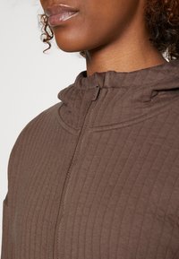 Sudadera con capucha marrón con cremallera y diseño nervado, que presenta un cuello alto, textura suave y una cremallera con una pequeña lengüeta.