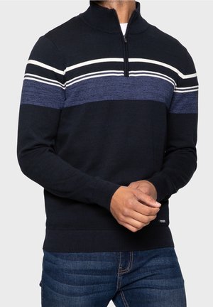 Gilet zippé navy avec des rayures horizontales blanches et bleues. Poignets et ourlet côtelés. Matière tricotée douce. Associé avec un jean bleu.