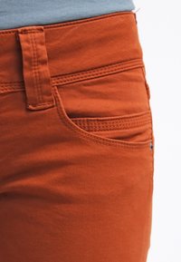 Pantalones de algodón naranja con frente plano, trabillas para cinturón y un bolsillo lateral con detalles de costura sutil. Textura suave.