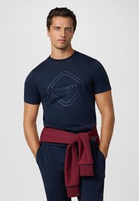 Marinblå t-shirt med en diamantlogotyp, bärs med en vinröd tröja knuten i midjan. Slät tyg med korta ärmar.