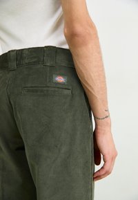 Groene corduroy broek met een patchzak en een merklabel op de achterkant, met een gestructureerd oppervlak en zichtbare naaidetails.