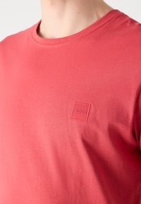 T-shirt rouge à col rond avec un petit logo carré en relief indiquant "BOSS" sur la poitrine gauche, porté par une personne à la peau claire.
