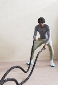 Black and white battle ropes are held by a person in a light gray long-sleeve top and light green pants, with light blue athletic shoes.