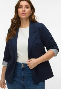 Navyblauwe blazer met opgerolde mouwen die een gestreepte binnenkant onthullen; gecombineerd met een wit topje en high-waisted spijkerbroek met dubbele knoopsluiting.