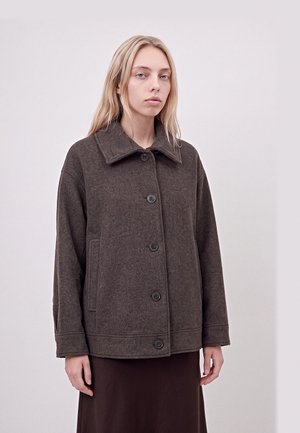 Un mannequin blond se tient devant un fond clair, vêtu d’une veste en laine brun foncé boutonnée sur le devant, à col large, et d’une longue jupe.