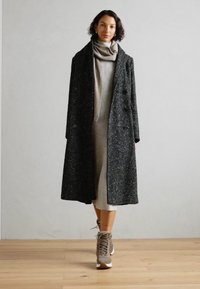 Long manteau gris foncé à la finition texturée, à double boutonnage, associé à une robe gris clair et des baskets beiges.