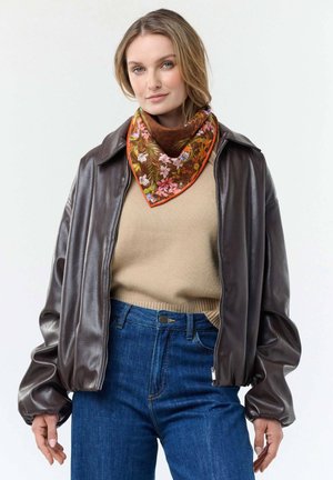 Woman wearing brown leather jacket, beige sweater, blue jeans, and a brown floral scarf with orange trim around her neck.