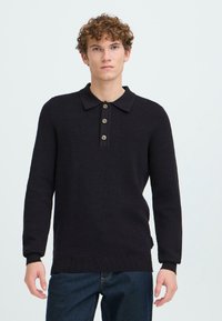 Mustassa neulotussa pitkähihaisessa polo-paidassa on kauluskera, jossa on neljä ruskeaa nappia sekä ribbiresorit hihansuissa ja helmassa.