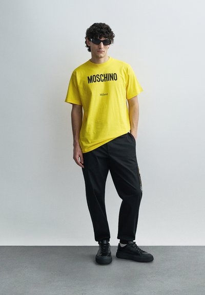 Sárga pamut póló vastag fekete "MOSCHINO" és "Milano" felirattal, laza fekete nadrággal és fekete sportcipővel párosítva, világos háttér előtt.