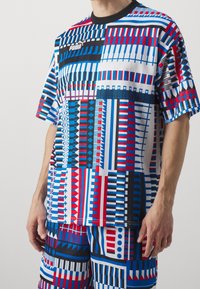 La camiseta PUMA multicolor presenta audaces patrones geométricos en rojo, azul y blanco, con un cuello negro y mangas cortas.
