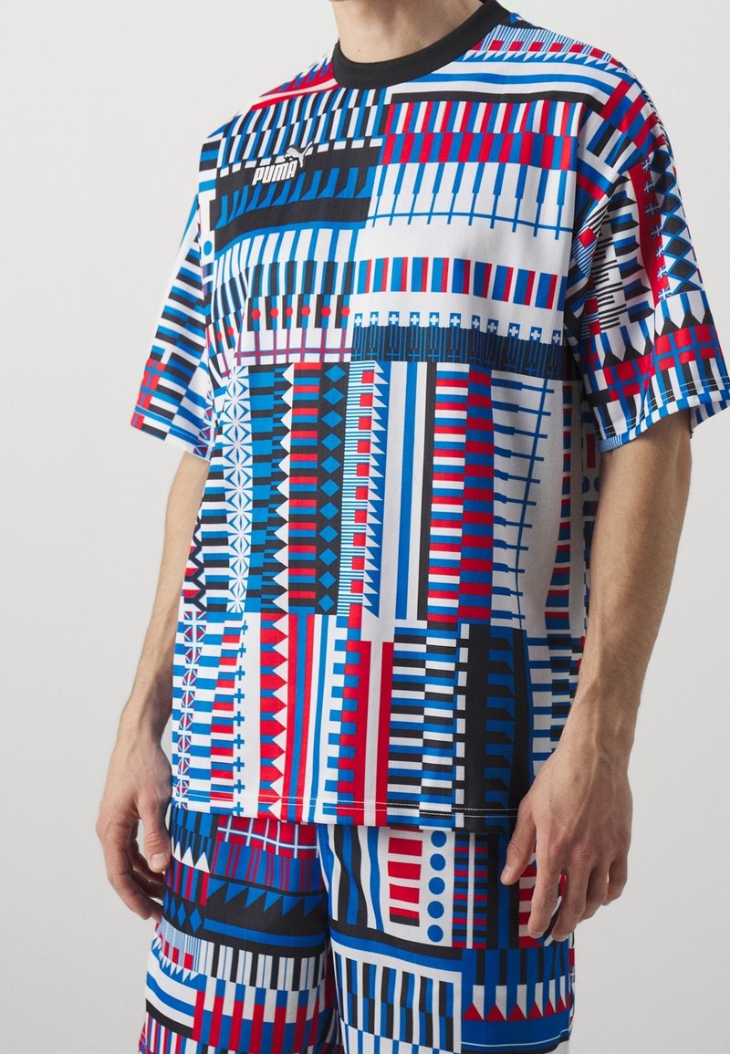 La camiseta PUMA multicolor presenta audaces patrones geométricos en rojo, azul y blanco, con un cuello negro y mangas cortas.