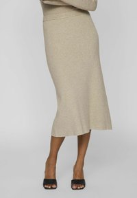 Jupe midi en tricot beige avec une légère évasement, présentant une texture côtelée et une finition lisse. Portée avec des talons ouverts noirs.
