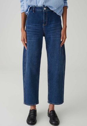 Person trägt gerade geschnittene blaue Jeans, schwarze Slipper und ein hellblau gestreiftes Hemd, das in die Hose gesteckt ist, und steht vor einem schlichten Hintergrund.