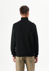 En person står med ryggen til, iført en sort sweatshirts og beige bukser, foran en helt hvid baggrund.