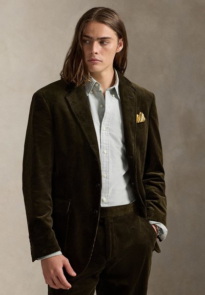 Σκούρο πράσινο σακάκι από κορδόνι με γιακά με εγκοπή, συνδυασμένο με ανοιχτό γαλάζιο πουκάμισο με κουμπιά και κίτρινο μαντηλάκι τσέπης με σχέδιο.