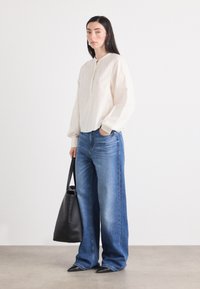 Una giovane donna si trova di fronte a uno sfondo bianco, indossando una blusa chiara e jeans larghi, mentre tiene in mano una borsa a tracolla nera.