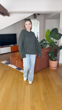 Übergroßer dunkelgrüner Pullover mit weiten Ärmeln, kombiniert mit hellblauen Schlagjeans und weißen Schuhen. Im Hintergrund ist eine Topfpflanze zu sehen.