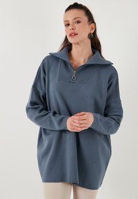 Gaiši zils adīts džemperis ar augstu apkakli, ar pusi rāvējslēdzēja aizdari, ribota tekstūra apkaklē un brīvas piedurknes.