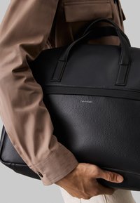 Sac de voyage noir en cuir avec une texture grainée, design horizontal et deux poignées supérieures. Présente le logo Calvin Klein à l'avant.