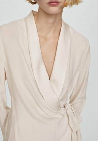 Krémové zavinovací šaty s hlubokým V-výstřihem, dlouhými rukávy, měkkou texturou a uvázaným detailem v pase, který zdůrazňuje draped design.