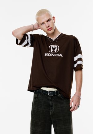 Brown short-sleeve t-shirt with white stripes and "HONDA" logo. Worn with dark patterned jeans and layered with a light-colored shirt.