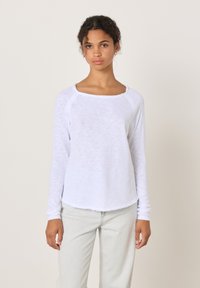 Junge Frau mit lockigem Haar, die ein weißes Langarmshirt und hellfarbene Jeans trägt, vor einem einfarbigen beigen Hintergrund stehend.