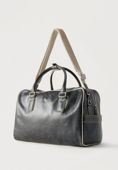 Sac de voyage en cuir noir avec une bandoulière beige réglable et deux petites poignées, forme rectangulaire, posé sur une surface blanche.