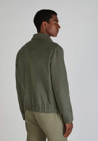 Grüne Fleecejacke mit Kragen, elastischem Saum und Nahtdetails, kombiniert mit hellgrünen Hosen, von hinten gesehen.