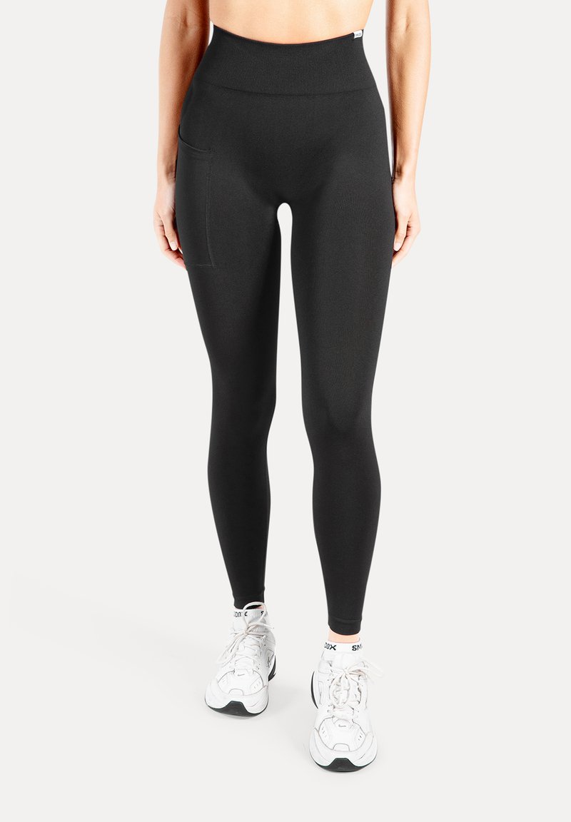 Schwarze Leggings mit hoher Taille aus elastischem Stoff, mit einer Seitentasche und glatter Textur, kombiniert mit weißen Sportschuhen.