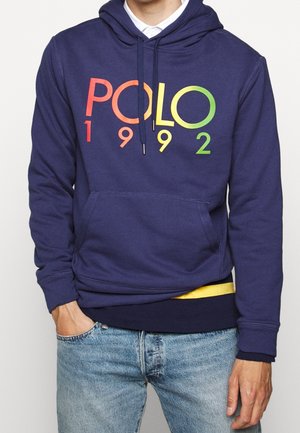 Homme portant un sweat à capuche bleu marine avec le texte « POLO 1992 » en dégradé rouge, jaune et vert, un jean en denim et une chemise jaune qui dépasse.