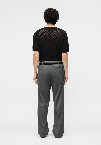 Άνδρας με σγουρά μαλλιά στέκεται γυρισμένος πλάτη, φορώντας μαύρο πλεκτό τοπ με κοντά μανίκια και χαλαρό γκρι παντελόνι με λεπτές ρίγες και μαύρη ζώνη.