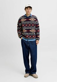 Jongeman staat in een gemêleerde wollen trui, denim shirt, losse donkere jeans, beige schoenen, witte pet en creolen tegen een effen achtergrond.