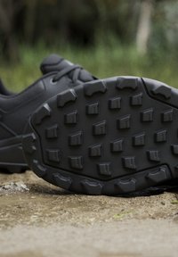 Suola nera di scarpa da trekking con profondi disegni del battistrada che poggia su un sentiero sterrato con fogliame verde sfocato sullo sfondo.