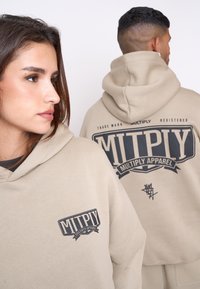 Felpa beige con logo anteriore che recita "MITPLY MULTIPLY APPAREL" e una grafica più grande sul retro. Presenta un cappuccio con coulisse e un tessuto morbido.