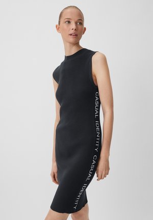 Schwarz gerippetes Strickkleid mit hohem Kragen und ärmellosem Design. Mit dem Text "CASUAL IDENTITY" vertikal an einer Seite.