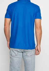 Modrá bavlněná polo košile s krátkým rukávem, plochým límcem a bočními rozparky, spárovaná s džíny v jemném odstínu. Jednoduchý design, bez vzorů.
