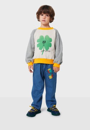 Bunter grauer Sweatshirt mit grünem Kleeblatt-Graphic, kombiniert mit blauen Hosen, die farbenfrohe Würfel zeigen. Schwarze und gelbe Schuhe mit strukturierten Sohlen.