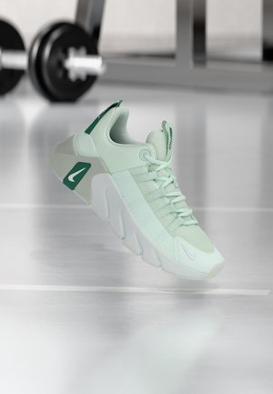 Lichtgroene sportschoen met dikke zool en veters, zwevend binnen met wazige fitnessgewichten op de achtergrond.