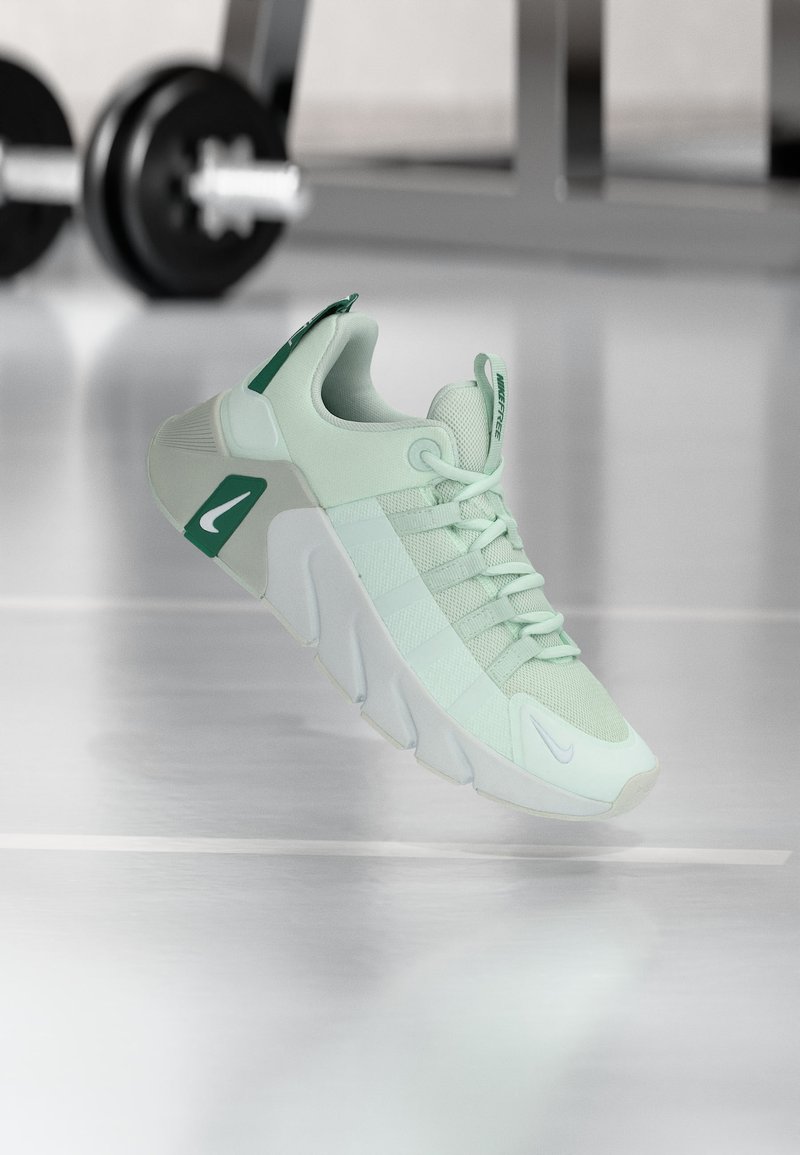 Αθλητικό παπούτσι ανοιχτού πράσινου με χοντρή σόλα και κορδόνια, αιωρούμενο μέσα σε κλειστό χώρο κοντά σε θολωμένα βάρη γυμναστηρίου στο παρασκήνιο.