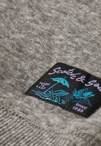 Grå strikket stof tæt på med et sort label med farverige grafik, herunder en strandparasol og planter, samt teksten "Made to Last."